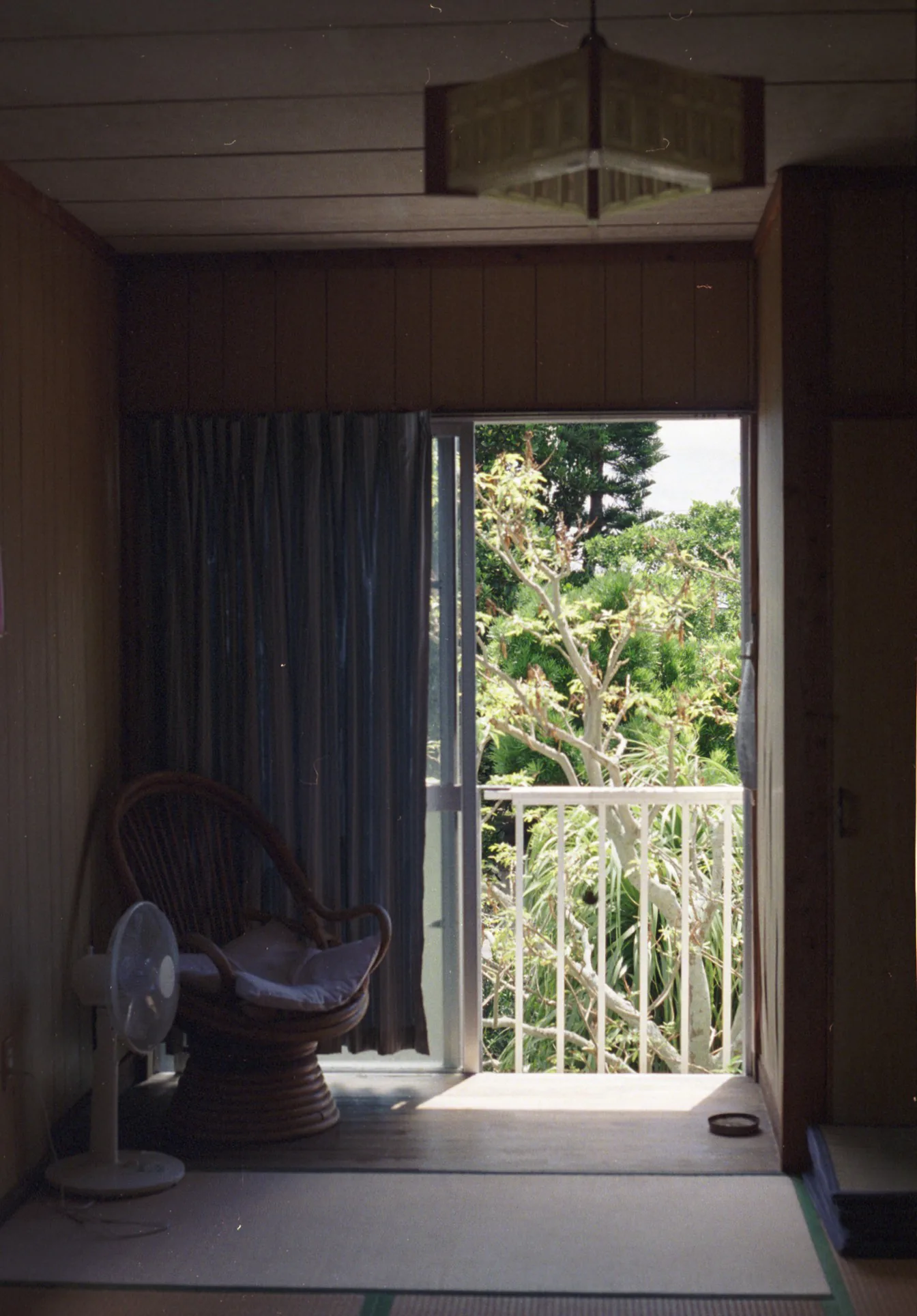 AI-generated alt text
English: Film photograph of a dim wooden room with tatami flooring, a rattan swivel chair and an electric fan beside a curtain. An open sliding door leads to a sunlit balcony with green trees beyond. Kuroshima, Okinawa, Japan.
日本語：畳敷きの薄暗い和室。カーテンのそばに籐の回転椅子と扇風機があり、開け放した戸の向こうに、日が差すバルコニーと庭木の緑が見える。沖縄県・黒島で撮ったフィルム写真。