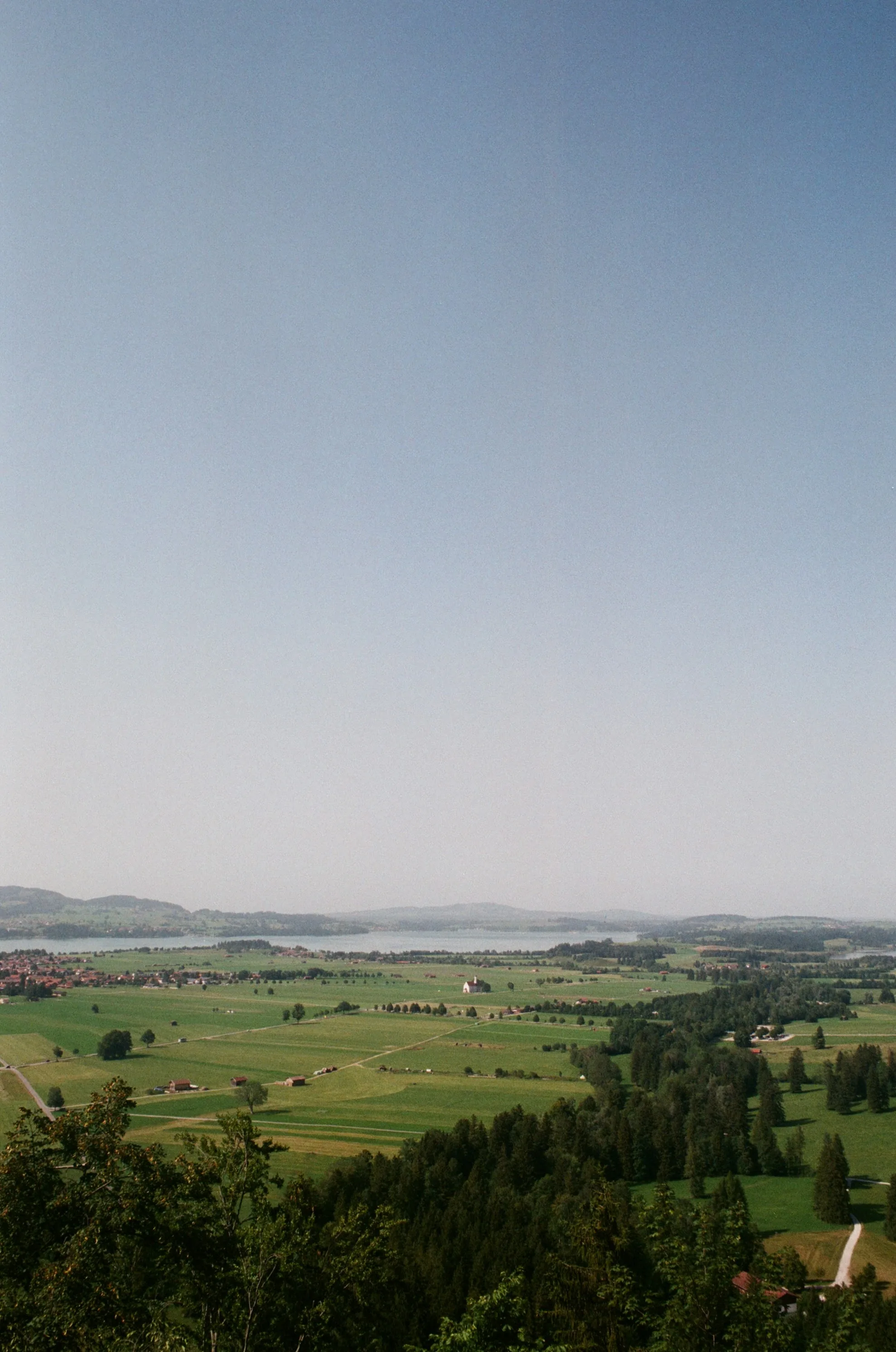 AI-generated alt text
English: Film photograph looking down from a hillside over green fields, farms and a lake toward distant hills, under a large hazy blue sky. Taken near Schwangau, Germany.
日本語：高台から見下ろした、緑の牧草地と点在する農家、その先に広がる湖と遠くの山並み。空は霞んだ青で、画面の大半を占めている。ドイツ・シュヴァンガウ近郊で撮ったフィルム写真。